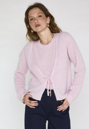 Femme portant un pull léger rose pâle et un cardigan noué à la taille, avec un pantalon bleu foncé, des boucles d'oreilles créoles en or et plusieurs bagues et bracelets en or.