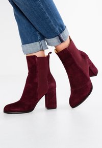 Stivaletti in suede bordeaux con tacco a blocco, dotati di pannelli laterali rigati e punta arrotondata. Indossati con jeans blu risvoltati.