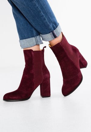 Bottines en daim bordeaux avec un talon empilé, dotées de panneaux latéraux ribbés et d'un orteil arrondi. Portées avec un jean bleu retroussé.
