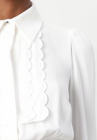 Blusa bianca con colletto, dettagli scallop lungo la pattina e maniche a sbuffo, realizzata in un tessuto leggero e liscio.