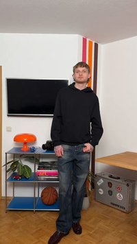 Tânăr îmbrăcat într-un hanorac negru și blugi stă într-o cameră cu un baschet, o cameră foto, o lampă portocalie și o valiză decorată cu abțibilduri.