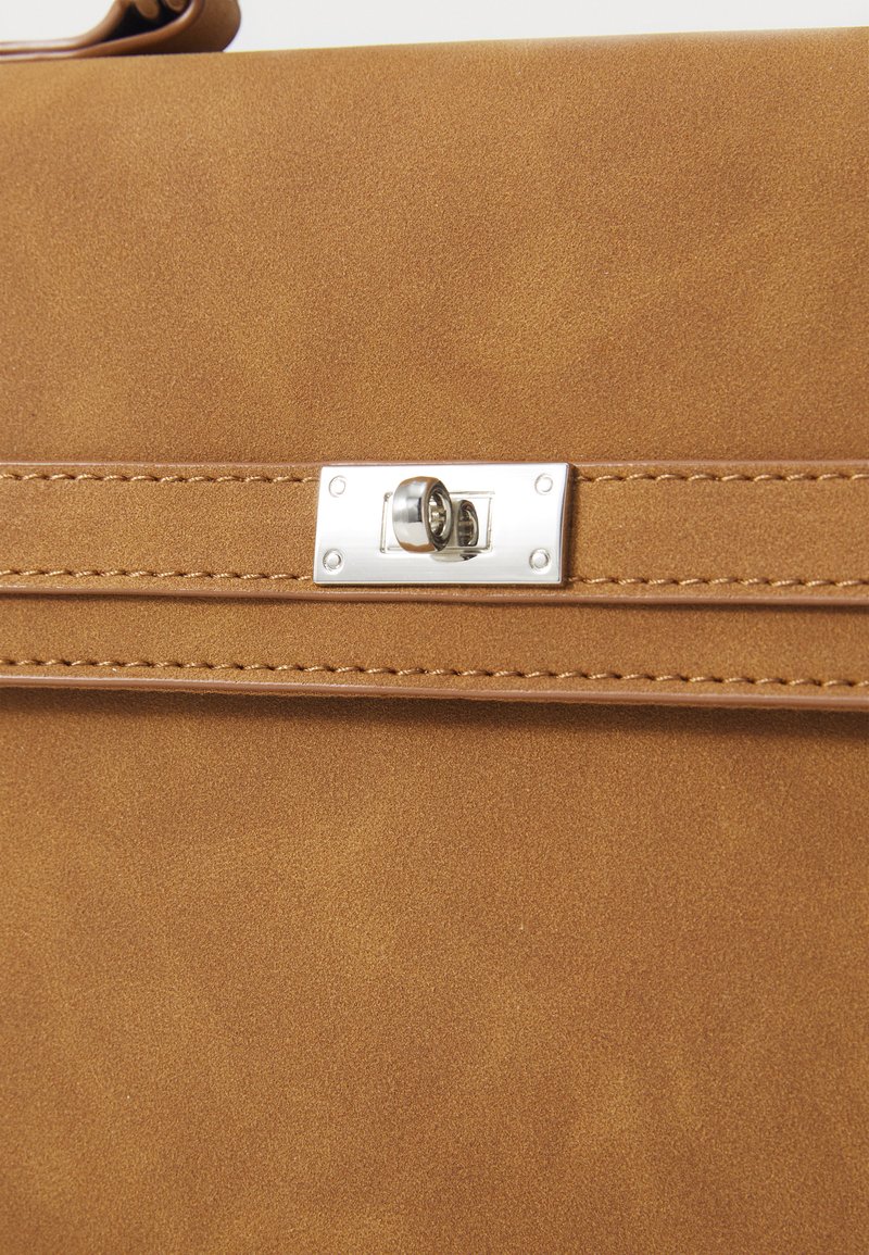 Szczegół brązowej torby z syntetycznej skóry z prostokątnym srebrnym zamkiem, kontrastowym szwem i gładką teksturą powierzchni.