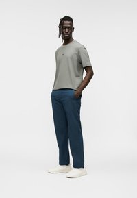 Άνδρας με πλεξούδες στα μαλλιά, ντυμένος με γκρι μπλουζάκι, μπλε παντελόνι και λευκά αθλητικά παπούτσια, στέκεται με τα χέρια στις τσέπες μπροστά σε απλό φόντο.