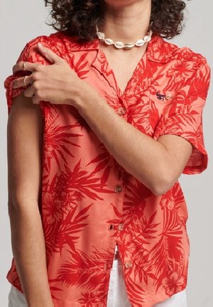 Osoba o kręconych włosach, ubrana w czerwoną koszulę z krótkim rękawem z tropikalnym wzorem i białym naszyjnikiem z muszli, poprawiająca lewy rękaw prawą ręką.