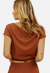 Crop top court de couleur rouille, avec une texture côtelée, un col bateau et un ourlet volanté. Présente une coupe décontractée et des détails subtils aux manches.