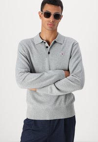 Pilkos ilgos rankovės polo marškiniai su prigludusia apykakle, trimis juodais mygtukais, gumuotais rankogaliais ir maža logotipo etikete ant kairės krūtinės.