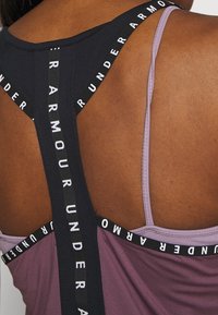 Sporttopp i svart och lavendel med bred svart axelrem som visar varumärkesnamnet i vitt, vilket framhäver en racerback-stil.