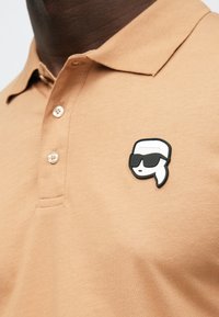 Barna poloing, galléros nyakkal, három gombbal és az bal mellkasrészen egy gumimatrica található, melyen napszemüveges, stilizált arc látható.