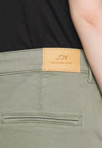 Olivgröna byxor med en texturerad läderetikett som har "JDY THE DENIM STORY" och en dubbelstickning längs midjan.