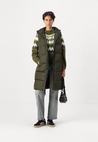 Lang olivengrønn puffer vest med hette, med glidelås foran og sidelommer, lagd over en mønstret strikkegenser og jeans.