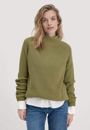 Olivengroene gebreide trui met een hoge hals en geribbelde textuur. Beschikt over lange mouwen en een relaxte pasvorm. Gedragen over een witte shirt met kraag.