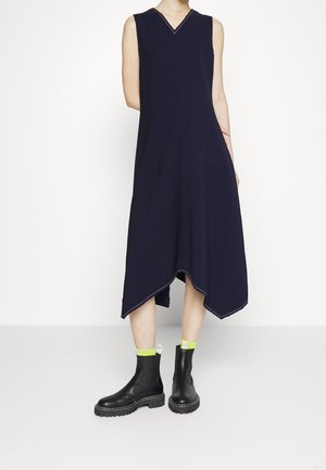 Persoon draagt een navy asymmetrische midi jurk zonder mouwen en zwarte chunky enkellaarsjes met neon groene sokken, staand tegen een witte achtergrond.