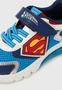 Sinivalkoinen lasten lenkkari, jossa siniset nauhat, Superman-logo sivulla ja tummansininen remmi, jossa teksti "Superman" ja Geox-brändäys.