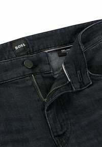 Dark denim jeans featuring a zip fly, button closure, and branded tag. The fabric has a smooth texture with minimal visible stitching.