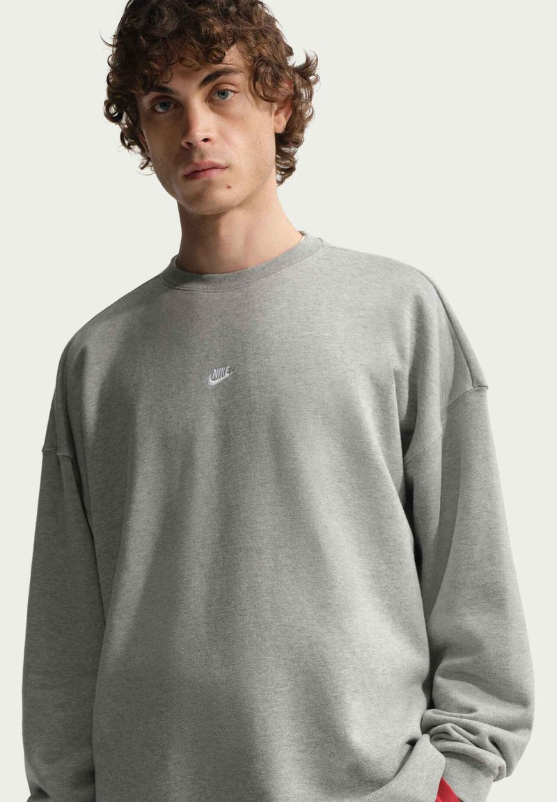 Jeune homme aux cheveux bouclés portant un sweat-shirt gris ample Nike sur un fond clair uni.