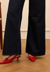 Jeans a gamba larga blu scuro con cuciture arancioni abbinate a mules con tacco rossi lucidi con un dettaglio a fiocco. Sfondo in legno.