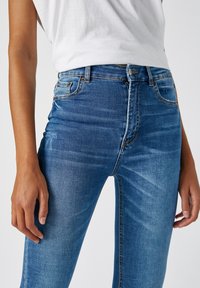 Pantalones vaqueros ajustados en denim azul medio, con cintura alta, un ligero desgaste, cinco bolsillos y detalles de costura tradicionales. Textura suave.