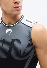 Κοντινό πλάνο ενός ατόμου που φοράει αμάνικο αθλητικό μπλουζάκι Venum σε γκρι και μαύρο, με λευκό λογότυπο φιδιού στο στήθος και επώνυμο πάνελ στον ώμο.