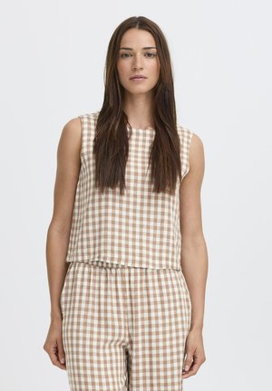 Frau mit langen braunen Haaren, die ein ärmelloses beiges und weiß kariertes Oberteil sowie passende Hosen vor einem einfarbigen Hintergrund trägt.