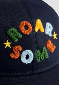 Tengerészeti sapka hímzett, többszínű betűkkel, amelyek a "ROAR SOME" feliratot formázzák, textúrált fonal kiemelésekkel és sárga csillag részletekkel.