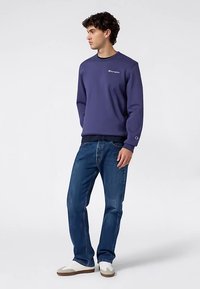 Granatowa bluza z haftowanym logo, ściągaczami na mankietach i dole, zestawiona z niebieskimi dżinsami i jasnymi trampkami.