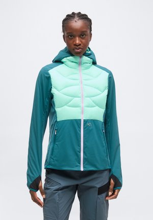 Femme portant une veste à capuche bleu sarcelle et vert menthe avec fermeture éclair et pantalon gris, debout devant un fond clair uni.