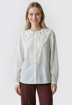 Giovane donna sorridente, indossa una camicetta bianca con bottoni e un grande colletto smerlato e pantaloni marrone scuro, in piedi su uno sfondo semplice.
