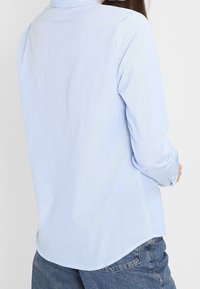 Vaaleansininen, raidallinen kauluksellinen ja pitkähihainen paita, jossa on kaareva helma. Valmistettu kevyestä, sileästä kankaasta.