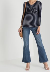 Marinblå och vit randig långärmad topp med inslaget design, kombinerad med utsvängda jeans i denim och vita klacksandaler, bärande en brun axelväska.