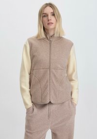 Hellbraune Fleece-Weste mit hohem Kragen und durchgehendem Reißverschluss; cremefarbene Ärmel und strukturiertes Material; seitliche Taschen und taillierter Schnitt.