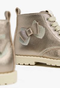 Metallisch goldene Knöchelstiefel mit strukturiertem Finish, beigen Schnürsenkeln und einem dekorativen Schleifenakzent an der Seite. Sie verfügen über eine Gummisohle für besseren Halt.