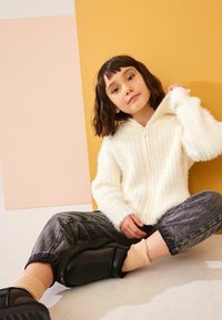 Felpa zip-up bianca a coste con texture morbida; jeans grigi larghi; calzature nere; calzini a coste beige; seduto contro uno sfondo colorato.