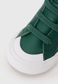 Zöld bőr sportcipő fehér gumitalppal, texturált fűzőrésszel, lekerekített orral és kontrasztos varrással.