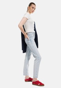 Jeans de tiro alto azul claro con un ajuste slim, llevados con una camiseta blanca de manga corta y zapatillas rojas, de pie contra un fondo liso.