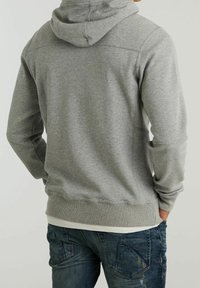 Hombre vestido con una sudadera gris lisa con capucha y jeans azules desgastados, mostrado de espaldas con las manos en los bolsillos.