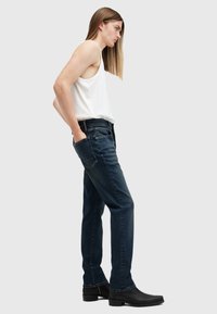 Jeans de mezclilla azul con corte recto y un ligero desgastado, combinados con una camiseta sin mangas blanca y botas negras hasta el tobillo sobre un fondo gris.
