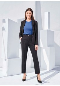 Schwarzes Blazer und maßgeschneiderte Hose, kombiniert mit einem blauen Oberteil. Modell trägt spitze schwarze High Heels. Geometrischer weißer Hintergrund.
