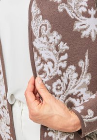 Cardigan marron à motifs avec un design floral blanc, en tissu tricoté texturé, à l'avant ouvert et avec des détails de bordure contrastants. Main tenant la manche.