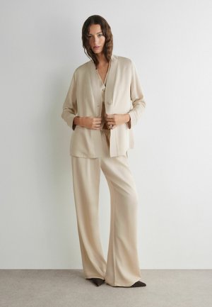 Frau passt einen beigen, knöpfbaren Blazer über passenden weiten Hosen an, steht vor einer schlichten weißen Wand auf grauem Boden.