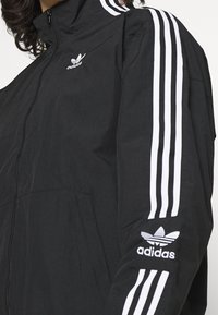 Svart zip-up jacka i lättviktsmaterial. Har vita trippelränder längs ärmarna och en broderad adidas-logga på bröstet och handleden.