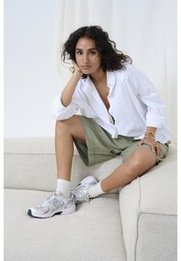 Chemise blanche à boutons, shorts vert olive et baskets New Balance grises avec des chaussettes blanches. Assis sur un canapé texturé de couleur crème.