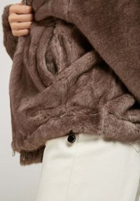 Veste en fausse fourrure de couleur marron douce avec une texture épaisse, dotée d'une fermeture éclair et de poches latérales discrètes. Pantalons de couleur crème visibles en dessous.