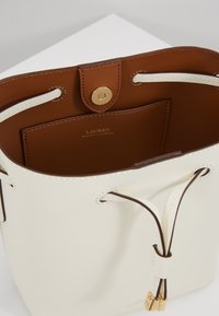Bílá kožená kabela ve tvaru kyblíku s hnědým interiérem, stahovací uzávěr, zlaté kování a logo "Lauren Ralph Lauren" uvnitř i zvenku.