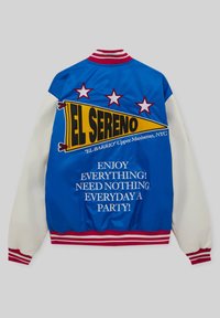 Veste universitaire bleu et blanc avec poignets rayés rouges, fanion "EL SERENO", trois étoiles, et texte faisant référence à Upper Manhattan, NYC, ainsi qu'un message de fête.