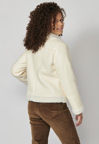 Krém színű műgyapjú dzseki texturált felülettel, széles gallérral és puha béléssel, barna kordbársony nadrággal párosítva.