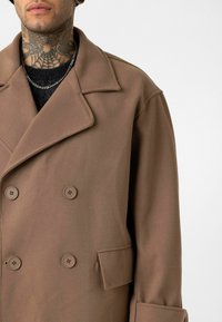 Man draagt een bruin double-breasted jas met brede revers, een zwarte trui, een zilveren ketting en een zichtbare nektattoo.