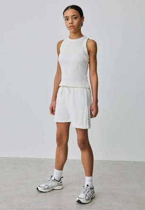 Noor naine seisab, kannab valget varrukateta topi, valge nööriga pükse, valgeid sokke ja valgeid tossusid halli põranda ja seinatausta ees.