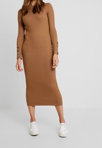 Abito midi a coste in marrone con maniche lunghe, caratterizzato da dettagli con bottoni sui polsini. Abbinato a sneakers bianche per un look casual.