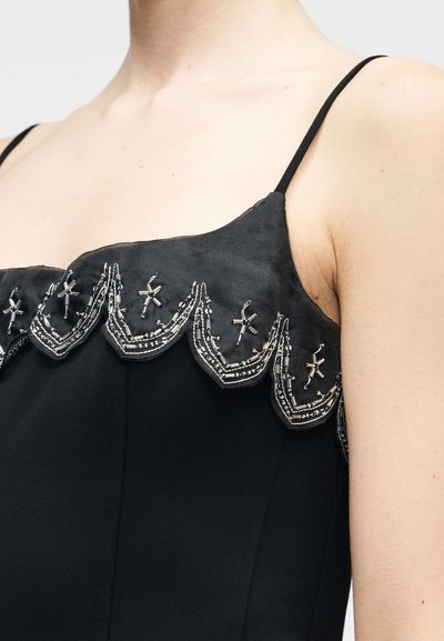 Melna kleita ar teksturētu kakla izgriezumu, izšuvu ar sarežģītiem sudraba pērlīšu rotājumiem un smalkām viļņveida malām. Plāni plecu siksniņas.