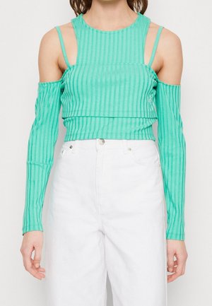 Femme portant un crop top côtelé bleu sarcelle à épaules dénudées avec des bretelles spaghetti et un pantalon blanc taille haute sur un fond uni.